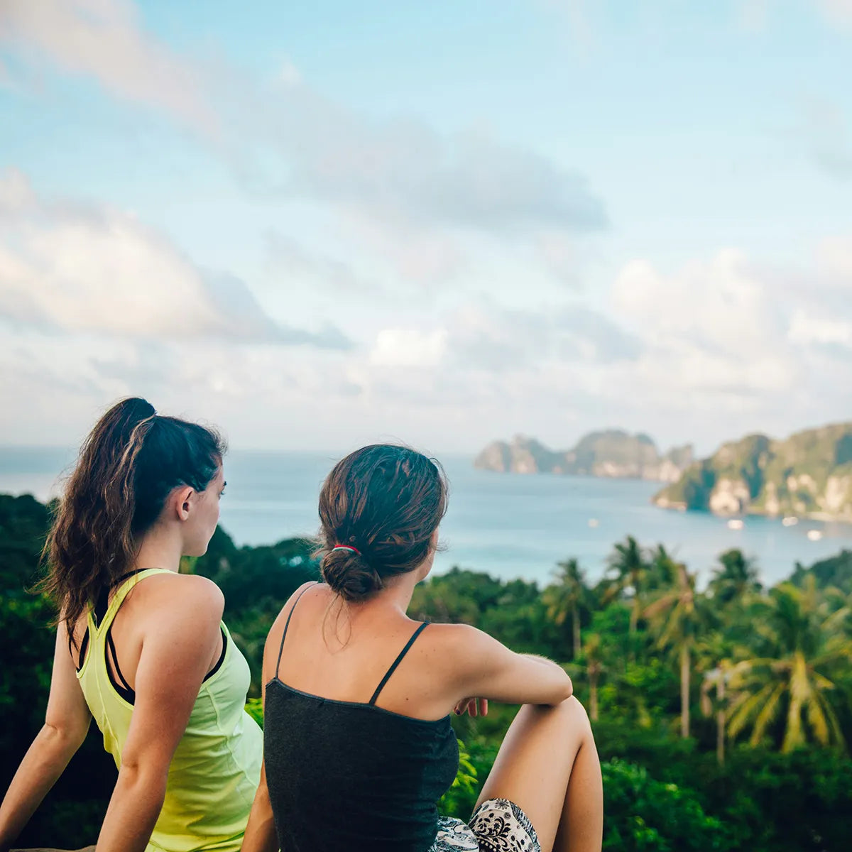 Two women sitting on a cliff overlooking a tropical landscape with ocean and mountains.