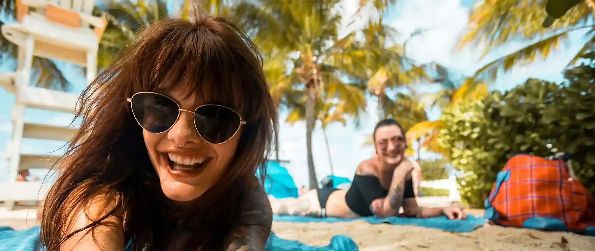 Woman wearing sunglasses and smiling on a beach with a man in the background.